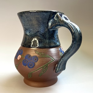 May include: A ceramic mug featuring a dark blue glazed upper section and a brown lower body. The brown area is decorated with a hand-painted design of blue berries on green stems. The mug includes a sturdy, matching dark blue curved handle.