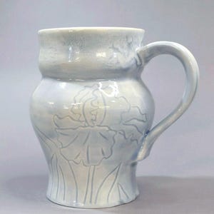 May include: A ceramic mug with a light blue and white glazed finish, featuring a subtle embossed floral design of an iris flower on the front. The mug has a rounded body, a wide rim, and a curved handle extending from the side of the vessel.