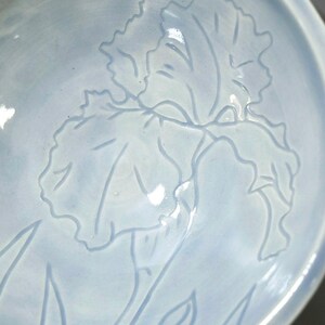 May include: A light blue ceramic bowl featuring a delicate, incised line drawing of an iris flower on its interior surface. The glaze has a smooth, glossy finish with subtle variations in color depth, highlighting the floral design.