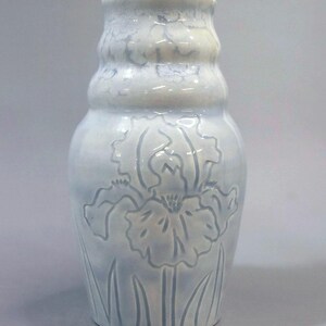 May include: A light blue ceramic vase with a glossy finish and a bulbous shape. The surface features a relief-molded iris flower design with long, slender leaves at the base. The neck of the vase has a subtle, undulating texture with a soft glaze.