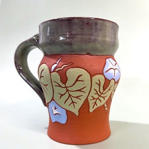 Peut inclure: Une tasse en céramique avec une base couleur terre cuite ornée de feuilles vert clair en relief et de fleurs de gloire du matin bleu pâle. La partie supérieure de la tasse et l'anse présentent une glaçure lisse, sombre, brillante et mouchetée gris-violet.