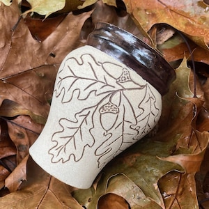 Puede incluir: Una taza de cerámica blanca con un borde marrón y un diseño de hojas de roble y bellotas. La taza está sentada sobre un lecho de hojas marrones.