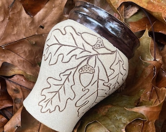 May include: A white ceramic mug with a brown rim and a design of oak leaves and acorns. The mug is sitting on a bed of brown leaves.