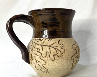 Könnte beinhalten: Eine braune und cremefarbene Keramiktasse mit Henkel. Die Tasse ist mit einem Muster aus Eichenblättern verziert.