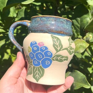 Pode incluir: Uma caneca de cerâmica com base na cor creme e borda e alça esmaltadas em azul. O corpo da caneca apresenta um design pintado à mão de um cacho de bagas azuis com folhas verdes. A caneca tem um formato arredondado e uma alça curva.