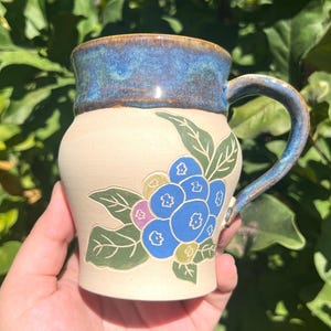 May include: A ceramic mug with a cream-colored base and a blue glazed rim and handle. The front features a hand-painted design of blue berries with green leaves and a small purple accent. The mug has a rounded body and a sturdy, curved handle.