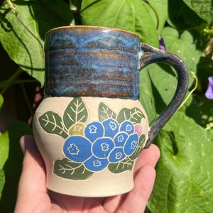 May include: A ceramic mug with a glossy blue glazed rim and handle, featuring a matte cream-colored base decorated with a hand-painted cluster of blue berries and green leaves. The mug has a cylindrical upper section and a wider, rounded lower body.