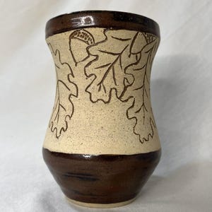 Peut inclure: Un vase en céramique avec un glaçage brun et crème. Le vase est décoré d'un motif en relief de feuilles de chêne et de glands.