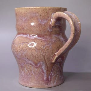 Peut inclure: Une tasse en céramique avec une silhouette incurvée et une anse unique. La surface présente une base beige mouchetée avec des émaux fluides et superposés dans des tons de rose poudré et de lavande douce, créant un aspect texturé et marbré.