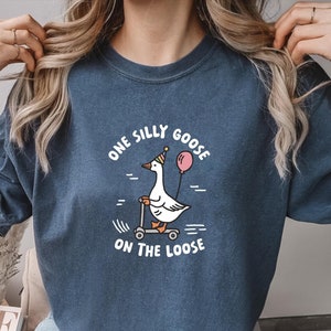 Puede incluir: Una camiseta azul marino con un gráfico blanco de un ganso con un gorro de fiesta y montando un scooter con un globo rosa. El texto de la camiseta dice "One Silly Goose On The Loose".