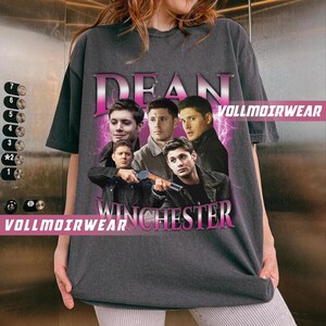 Puede incluir: Una camiseta gris oscura con un estampado gr&aacute;fico rosa de las palabras "Dean Winchester" e im&aacute;genes de cuatro hombres. El estampado gr&aacute;fico tiene una est&eacute;tica vintage.