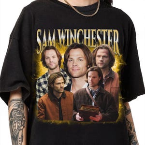 Puede incluir: Camiseta negra con un estampado gráfico de Sam Winchester de la serie de televisión Supernatural. El gráfico presenta cuatro imágenes de Sam Winchester en diferentes poses, con el texto "Sam Winchester" en letras doradas grandes en la parte superior.