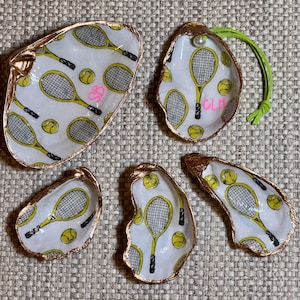 May include: Five gold-rimmed oyster shell dishes with a white background and a tennis racket and tennis ball pattern. The dishes are decorated with black tennis rackets and yellow tennis balls. One dish has the initials "CLA" written in pink.
