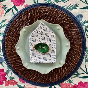 May include: A place setting with a blue and white patterned napkin, a green and gold oyster shell place card holder with the name "Claire" written on it, and a light blue plate on top of a brown woven charger.