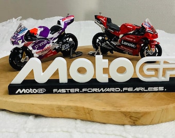 以下が含まれることがあります： 木製の台座に、紫と白、赤の2台のミニチュアMotoGPレーシングバイクが置かれています。 白い「MotoGP」ロゴが中央に配置され、その下に「FASTER.FORWARD.FEARLESS.」の文字があります。