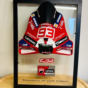 Può includere: Un espositore incorniciato che presenta una carenatura da moto rossa, bianca e blu con il numero 93, Monster Energy, Lenovo e altri loghi di sponsor. Sotto la carenatura, una targa riporta 1926-2026, Ducati Lenovo Factory Team e Desmosedici GP 2026 Marquez.