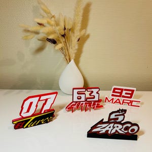 May include: A selection of red, white, and black racing-themed table decorations. Each decoration displays a number and a name, with a white vase and dried flowers in the background.