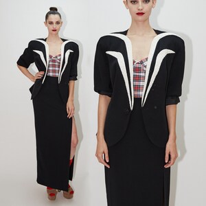 May include: Two women wearing black blazers with white accents and black skirts. The blazers have a unique design with a V-shaped neckline and white trim. The skirts are long and have a slit on the side.