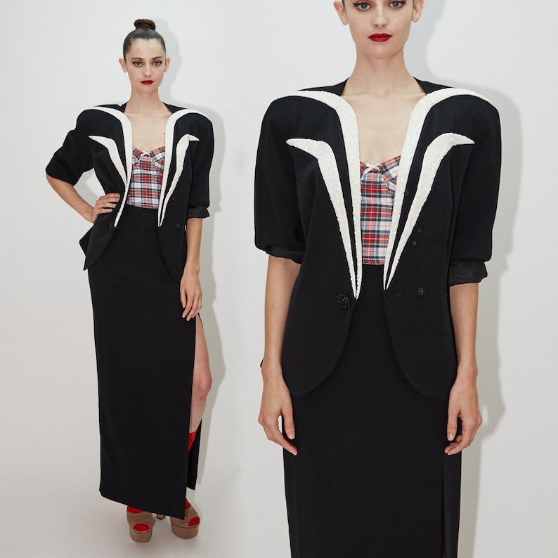 May include: Two women wearing black blazers with white accents and black skirts. The blazers have a unique design with a V-shaped neckline and white trim. The skirts are long and have a slit on the side.