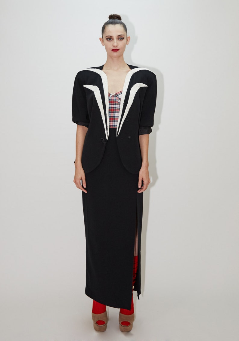 May include: A black blazer with a white collar and a white design on the front. The blazer is worn over a red and white plaid shirt and a black skirt with a slit up the side. The person is wearing red shoes.