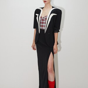May include: A woman wearing a black blazer with a white collar and a red and black plaid top. She is wearing a black skirt with a slit and red socks. She is wearing black heels.