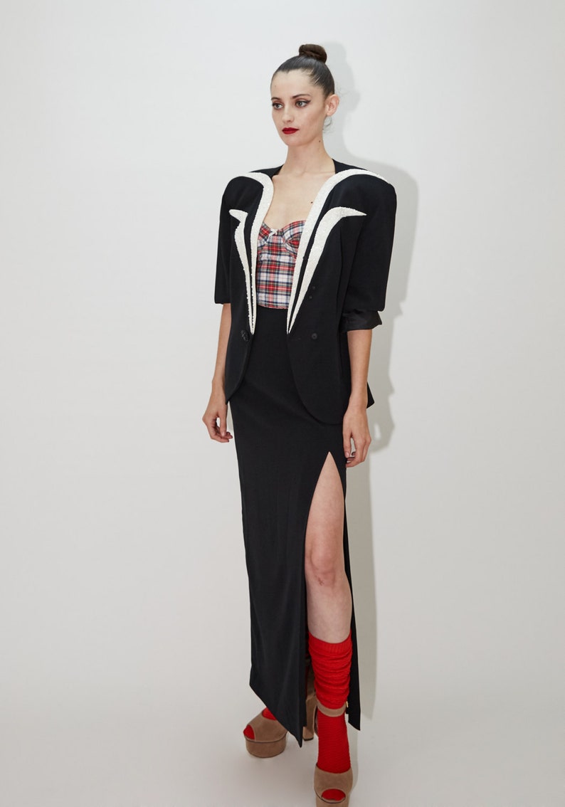 May include: A woman wearing a black blazer with a white collar and a red and black plaid top. She is wearing a black skirt with a slit and red socks. She is wearing black heels.