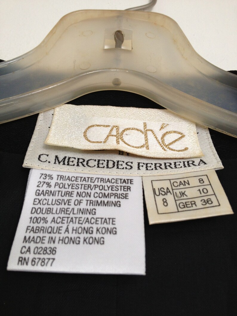 May include: White care label with black text detailing the fabric composition and care instructions for a black garment. The label reads "73% Triacetate/Triacetate 27% Polyester/Polyester Garniture Non Comprise Exclusive of Trimming Doublure/Lining 100% Acetate/Acetate Fabrique &agrave; Hong Kong Made in Hong Kong CA 02836 RN 67877". A smaller white label with black text shows size conversions for the garment: "USA 8 CAN 8 UK 10 GER 36".