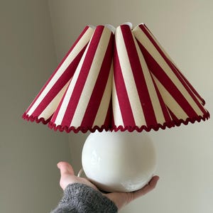 Pode incluir: Uma luminária de mesa de cerâmica branca com base redonda e abajur listrado. O abajur apresenta listras vermelhas e creme alternadas e uma borda ondulada. A luminária é segurada em uma mão.