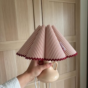 May include: A small table lamp with a cream-coloured base and a red and white striped lampshade. The lampshade has a scalloped edge and the base has a handle. The lamp is plugged in with a white cord.