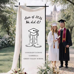 May include: A graduation scene featuring a banner with the text "class of 2026 He did it!" and "Congratulations Gabriella Green Valley School." Two graduates, one in white and one in black gowns, pose in front of the banner. Floral displays and mortarboards are arranged on the ground.