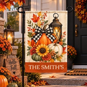 May include: A rectangular garden flag featuring a central illustration of orange and white pumpkins, a sunflower, autumn leaves, berries, and a black lantern with a black and white buffalo check bow. The text THE SMITH'S is printed in white.