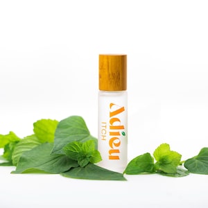 May include: A clear glass roll-on bottle with a wooden cap and a white label that says "Adieu Itch". The bottle is surrounded by green leaves.
