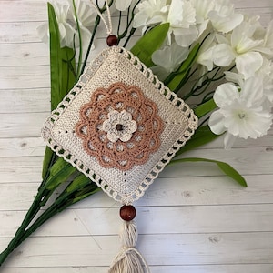 Könnte beinhalten: Ein quadratischer, cremefarbener Wandbehang aus Häkelarbeit mit einem Blumendesign in Braun- und Cremetönen. Er hat eine dekorative Quaste und eine Holzperle. Das Stück wird vor einem Hintergrund aus weißen Blumen und grünen Blättern präsentiert.