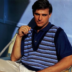May include: A man wearing a blue and white striped knitted vest over a navy blue polo shirt. He is sitting with his arm resting on his knee.