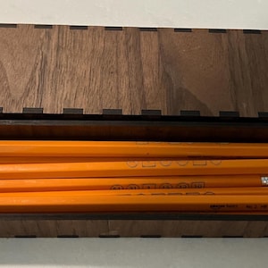 May include: A wooden pencil box with a hinged lid, containing several bright yellow pencils with pink erasers. The box and pencils are set against a light background.