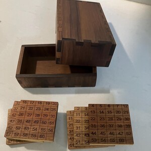 May include: A wooden puzzle box with an open lid, revealing a compartment and several wooden tiles. The tiles are engraved with numbers in a grid pattern. The box and tiles are made of dark wood.