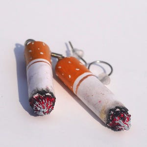 Op de afbeelding: Een paar novelty oorbellen in de vorm van aangestoken sigaretten. Elke oorbel heeft een witte cilinder met een oranje punt en een rood-zwart gloeddetail, bevestigd aan een zilverkleurige oorhaak.