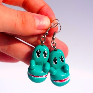 Könnte beinhalten: Ein Paar türkisfarbene Modeschmuck-Ohrringe. Jeder Ohrring zeigt eine cartoonartige Figur mit schwarzen Wimpern, weißen Augen und einem rot-weiß gestreiften Mund. Die Ohrringe hängen an silberfarbenen Haken.
