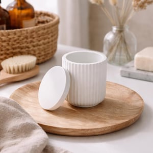 Könnte beinhalten: Ein weißer Keramikbehälter mit Deckel und vertikalen Rippendetails steht auf einem runden Holztablett. Der Behälter befindet sich neben seinem Deckel. Die Szene umfasst eine Holzbürste, Seife und einen geflochtenen Korb mit braunen Flaschen.
