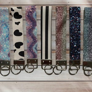 Puede incluir: Una colección de siete llaveros de muñeca con varios diseños. Los llaveros presentan patrones como purpurina, estampado de vaca, morado ondulado, rayas blancas y negras y purpurina sólida. Cada llavero tiene un cierre de metal plateado y un llavero.