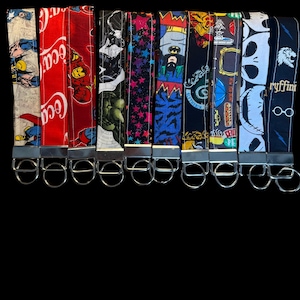 Puede incluir: Una colección de llaveros de tela con varios diseños, incluyendo Superman, Coca-Cola, Batman y patrones de estrellas. Cada llavero está unido a un anillo de metal plateado. El fondo es negro liso.