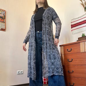 May include: A long, sheer duster in a black and white floral pattern. The duster is worn over a black top and blue jeans. The duster has long sleeves and a flowing silhouette, creating a layered look. The overall style is bohemian and casual.