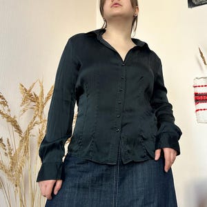 Op de afbeelding: Een zwarte blouse met knopen en lange mouwen. De blouse heeft een kraag en een getailleerde pasvorm. De stof lijkt een glad, mogelijk satijnachtig materiaal te zijn. De blouse wordt gecombineerd met een donkere spijkerbroekrok.