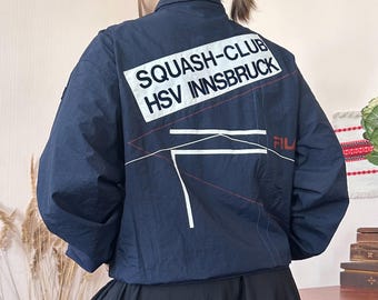 May include: A navy blue windbreaker jacket featuring a white rectangular patch on the back with the black text SQUASH-CLUB HSV INNSBRUCK. The jacket includes thin red and white geometric line accents and a small red FILA logo on the back.