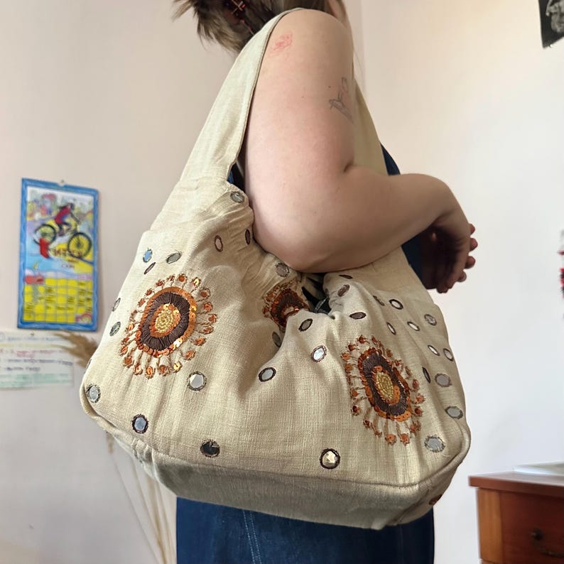 May include: A beige hobo bag featuring circular embroidered patterns in shades of brown and gold, accented with small reflective mirror discs scattered across the fabric surface. The bag has a wide shoulder strap and a soft, unstructured shape.