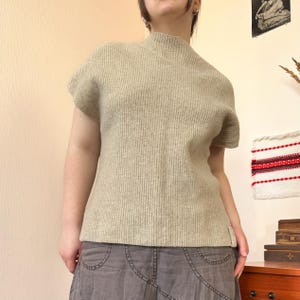 Puede incluir: Un suéter beige claro de manga corta con textura acanalada y cuello alto. El suéter está hecho de un material suave y tejido. La modelo lleva una falda gris.