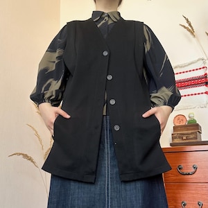 Könnte beinhalten: Eine schwarze, lange Weste mit Knopfleiste und Taschen. Die Weste wird über einer gemusterten Bluse mit langen Ärmeln getragen. Die Bluse hat eine Mischung aus olivgrünen und schwarzen abstrakten Formen. Das Outfit wird mit einem Jeansrock kombiniert.