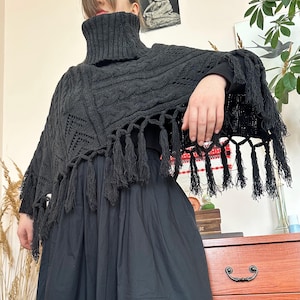 Op de afbeelding: Een zwarte gebreide poncho met een hoge coltrui en franjes. De poncho heeft een kabelgebreid ontwerp aan de voorkant en een decoratief opengewerkt patroon. De franjes hangen langs de onderkant van de poncho.