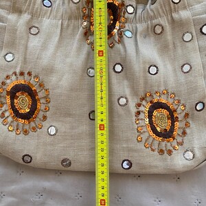 May include: A beige fabric shoulder bag decorated with small circular mirrors and embroidered floral motifs in brown, gold, and orange sequins. A yellow measuring tape is placed vertically over the center, showing a length of approximately 10 inches.