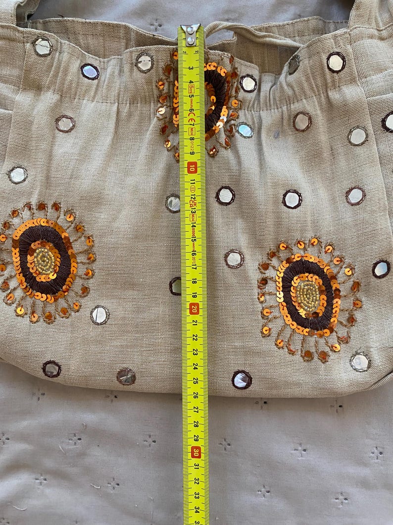 May include: A beige fabric shoulder bag decorated with small circular mirrors and embroidered floral motifs in brown, gold, and orange sequins. A yellow measuring tape is placed vertically over the center, showing a length of approximately 10 inches.